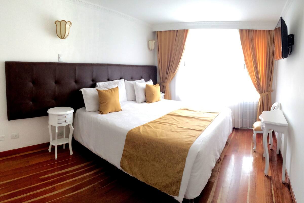 A hotel room with a large bed, a bedside table, a small white chair, and a television mounted on the wall.