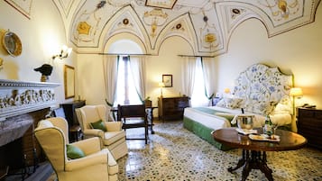 Habitación de hotel con chimenea, cama con cabecera floral, mesa de comedor con jarra y copa de vino, y candelabro.