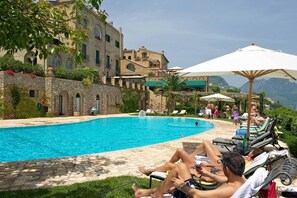 A poolside area with lounge chairs and umbrellas, a stone wall, and a building with arched windows and balconies. - Villa Cimbrone (Ravello)