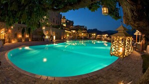 Una piscina iluminada con borde de piedra, rodeada de árboles y muros de piedra, con un edificio al fondo.