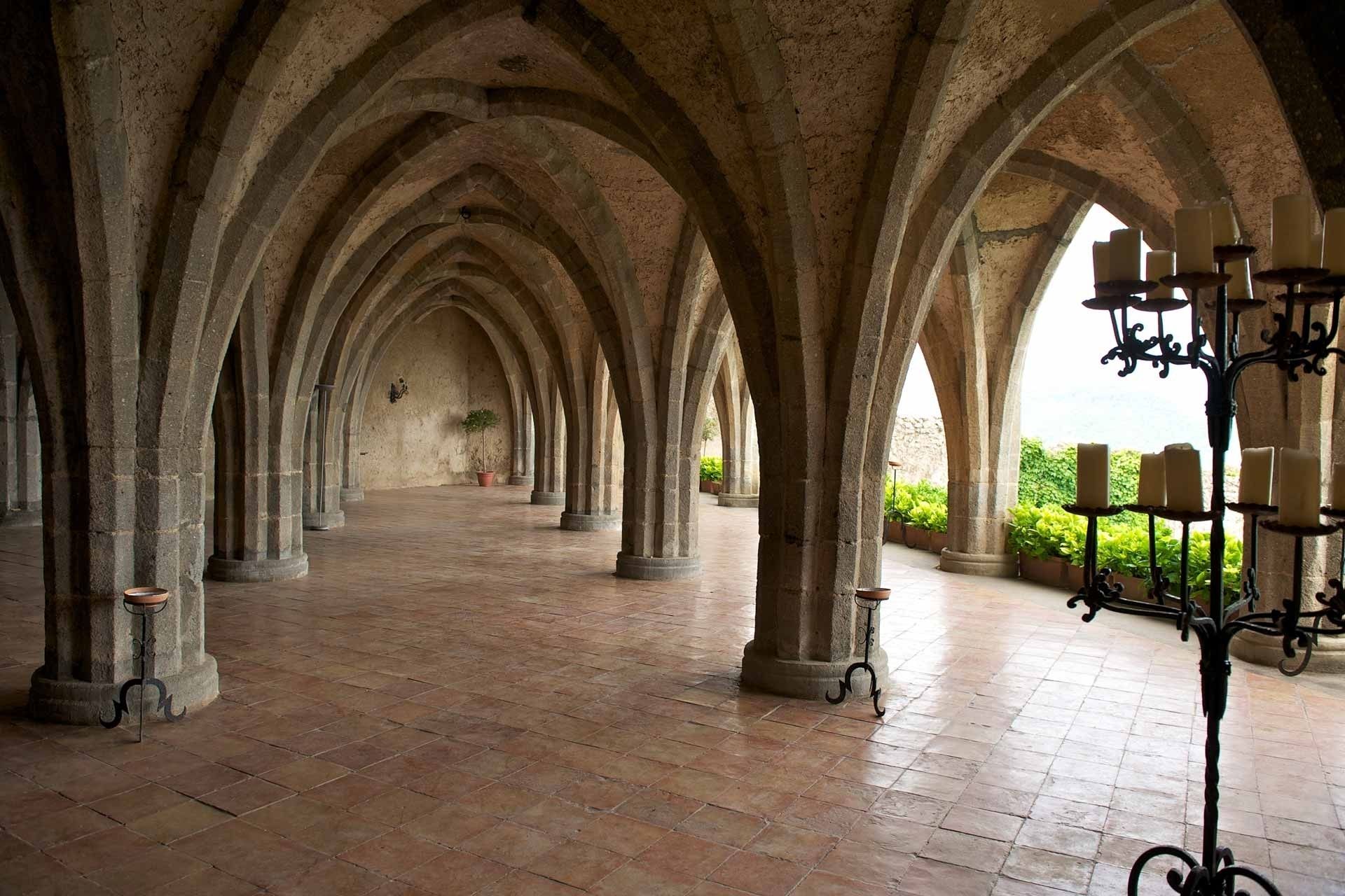 Un couloir bordé d’arches en pierre et doté d’un sol carrelé, qui mène à un espace ouvert donnant sur un jardin.