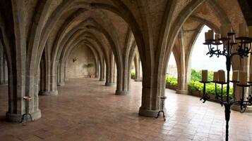 Un couloir bordé d’arches en pierre et doté d’un sol carrelé, qui mène à un espace ouvert donnant sur un jardin.