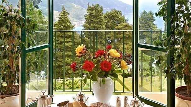 Una mesa de desayuno preparada con té, café, pastelería y fruta, con vistas a montañas a través de un amplio ventanal de arco.