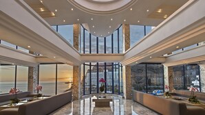 Eine geräumige Lobby mit hoher Decke, großen Fenstern mit Meerblick und einer zentralen Sitzecke mit Marmorboden.