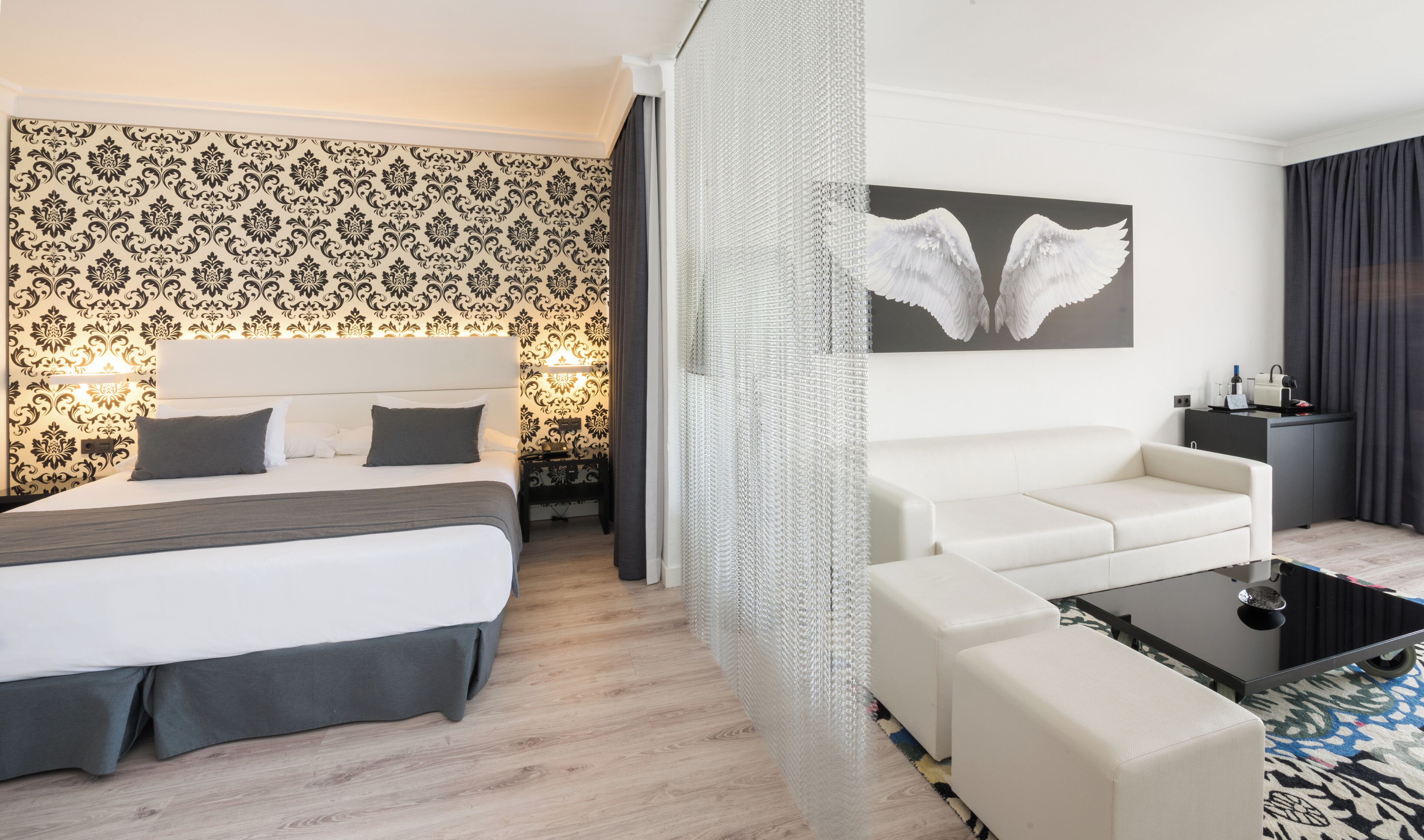 Hotellihuone, jossa on sänky, sohva, sohvapöytä, koristeellisella kuviolla varustettu seinä ja siipiaiheinen maalaus.