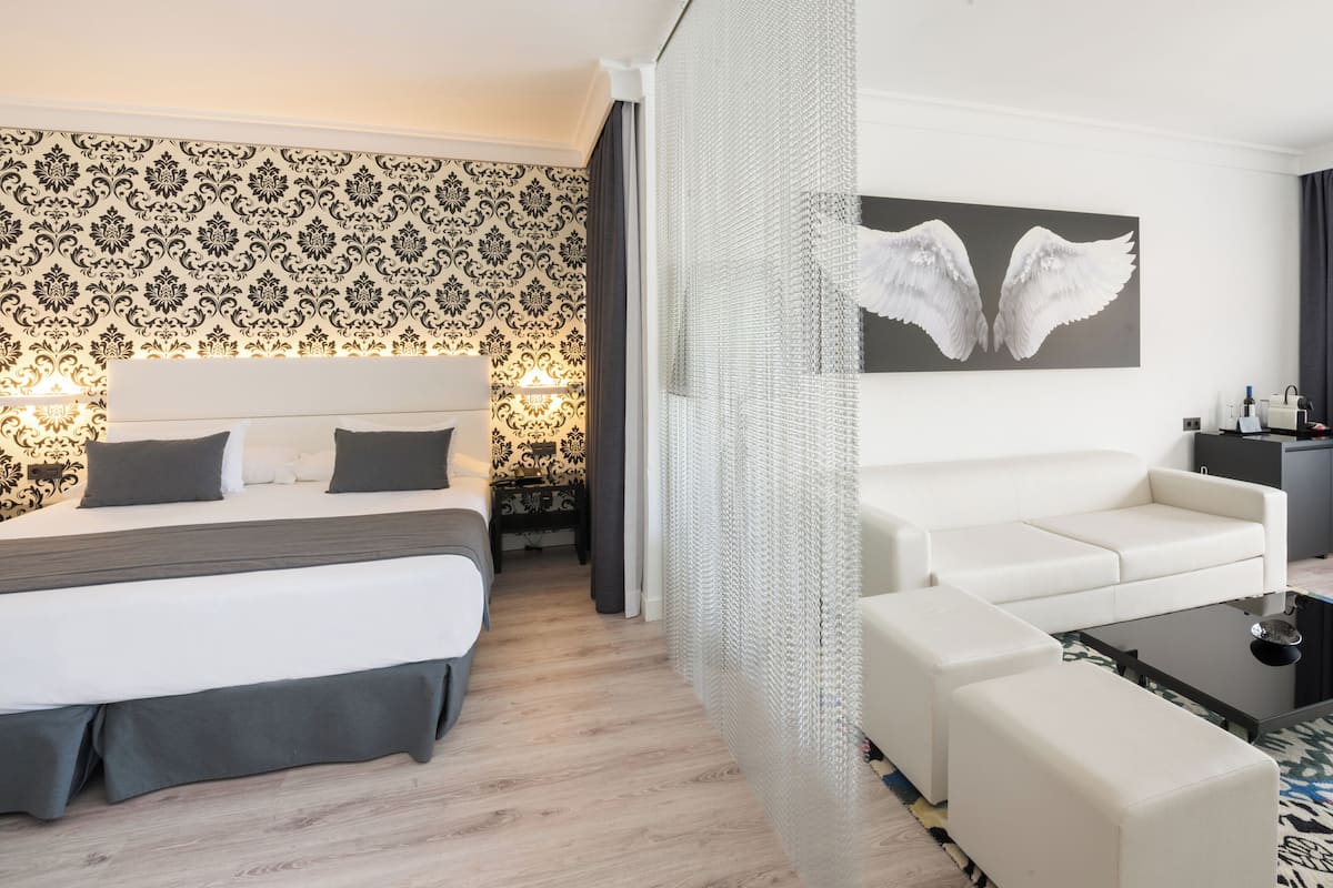 Hotellihuone, jossa on sänky, sohva, sohvapöytä, koristeellisella kuviolla varustettu seinä ja siipiaiheinen maalaus.