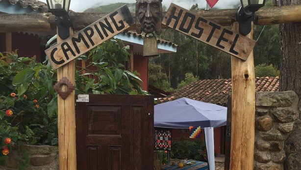 A wooden archway with "CAMPING HOSTEL" signage, a closed wooden door, and a stone wall. - Mystical Adventures Hostel (Urubamba)