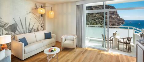A modern living room with a sofa, a small table, and a chair. There is a balcony with a view of the ocean and cliffs.