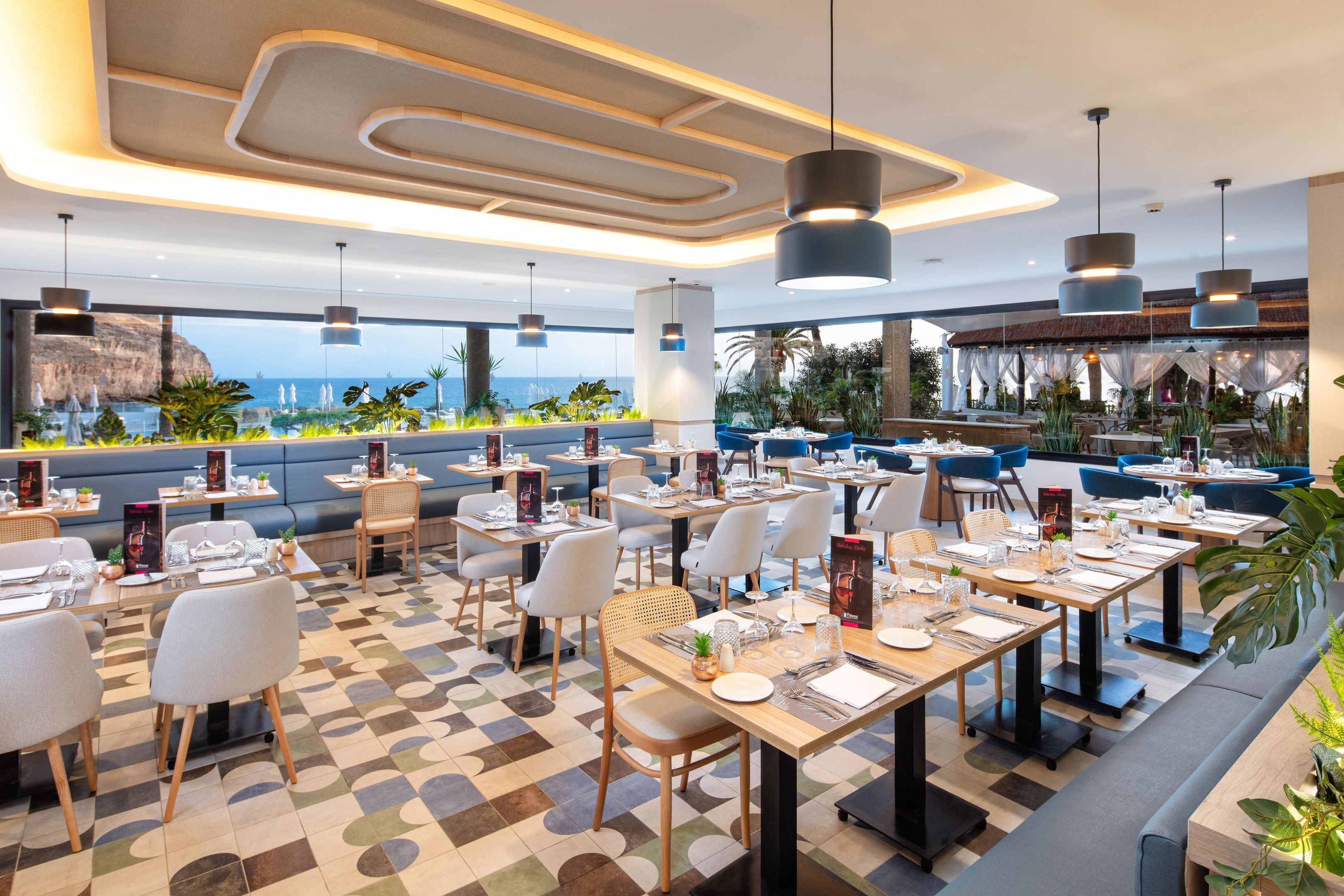 A modern restaurant with tables set for dining, large windows overlooking the sea, and a variety of plants and greenery.