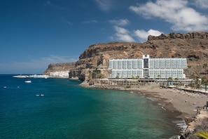 A large hotel with multiple floors, located near a beach with clear blue waters and rocky cliffs. - Hotel Princess Taurito - All Inclusive (Mogan)