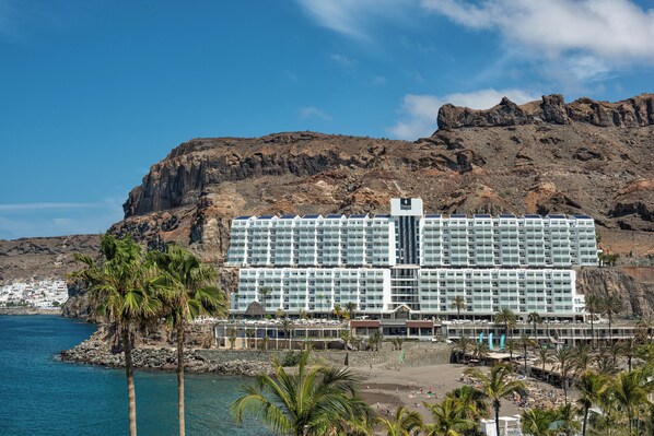 A large hotel with multiple floors, a beach in the foreground, and a rocky cliff in the background. - Hotel Princess Taurito - All Inclusive (Mogan)