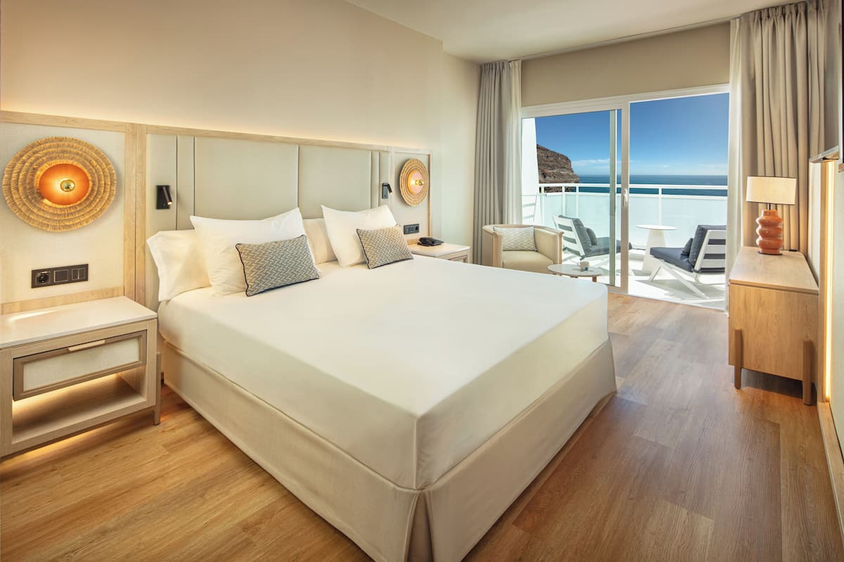 A hotel room with a large bed, a nightstand, a chair, a table, and a view of the sea.