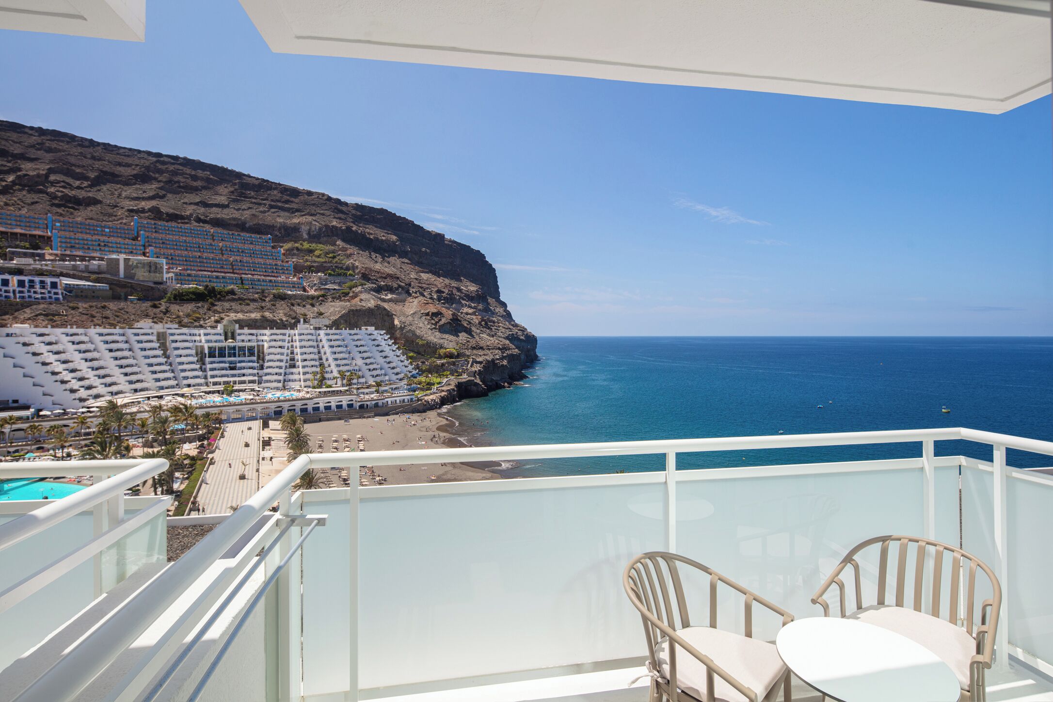A balcony with a view of a beach and ocean, white chairs, and a pool.