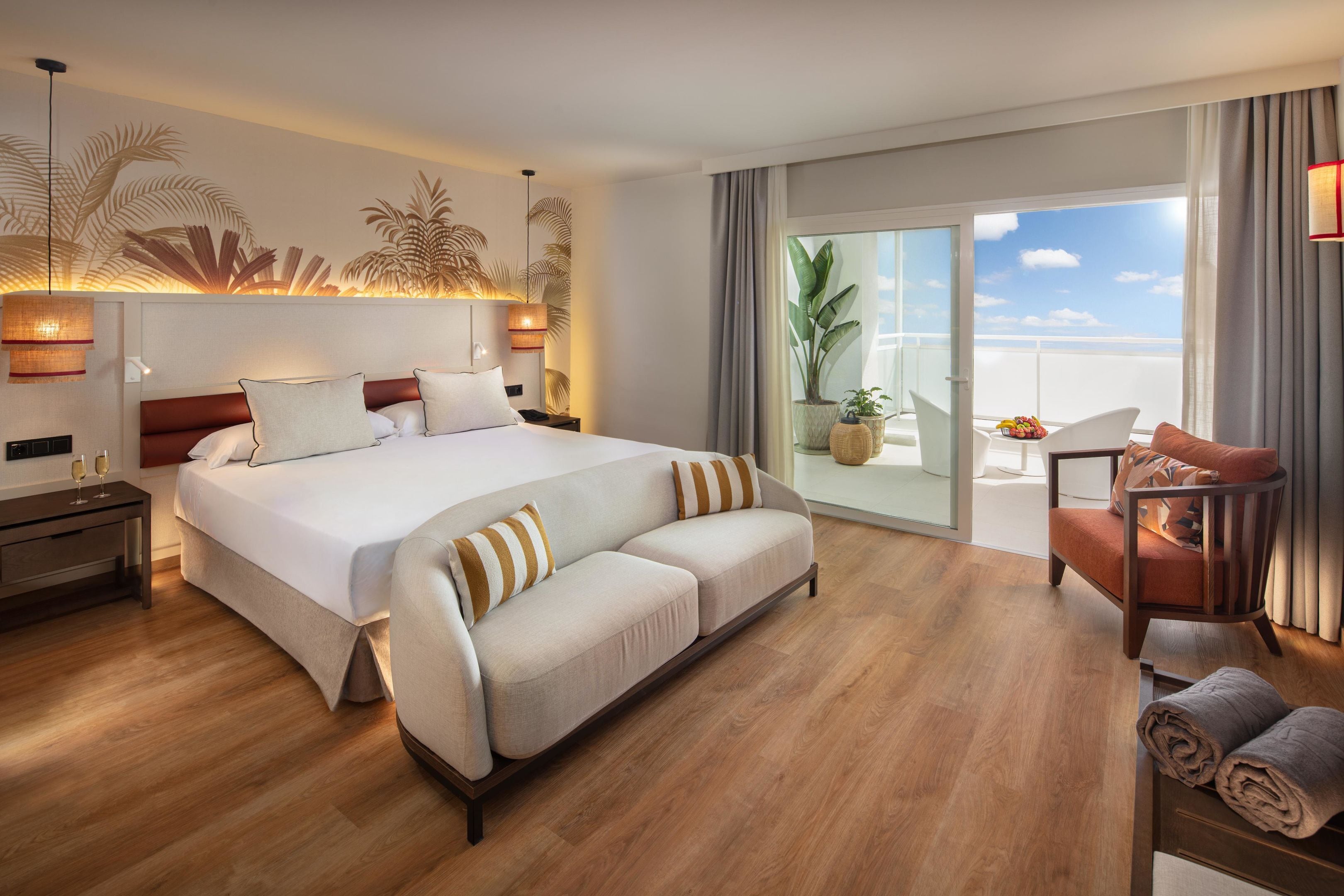 A modern hotel room with a large bed, a sofa, a chair, and a balcony with a view.