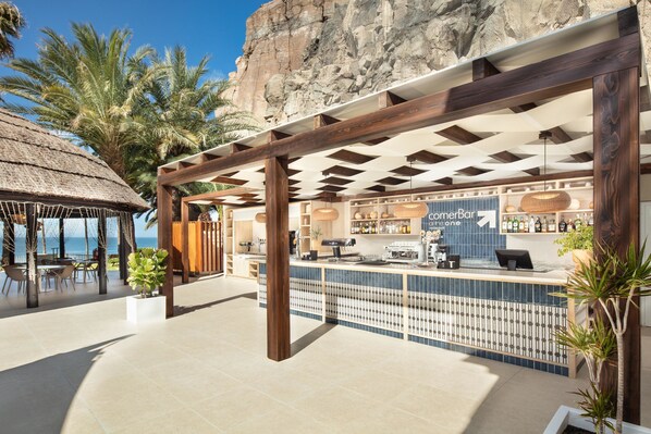 A beachside bar with a thatched roof, a counter with a blue sign, and palm trees. - Hotel Princess Taurito - All Inclusive (Mogan)