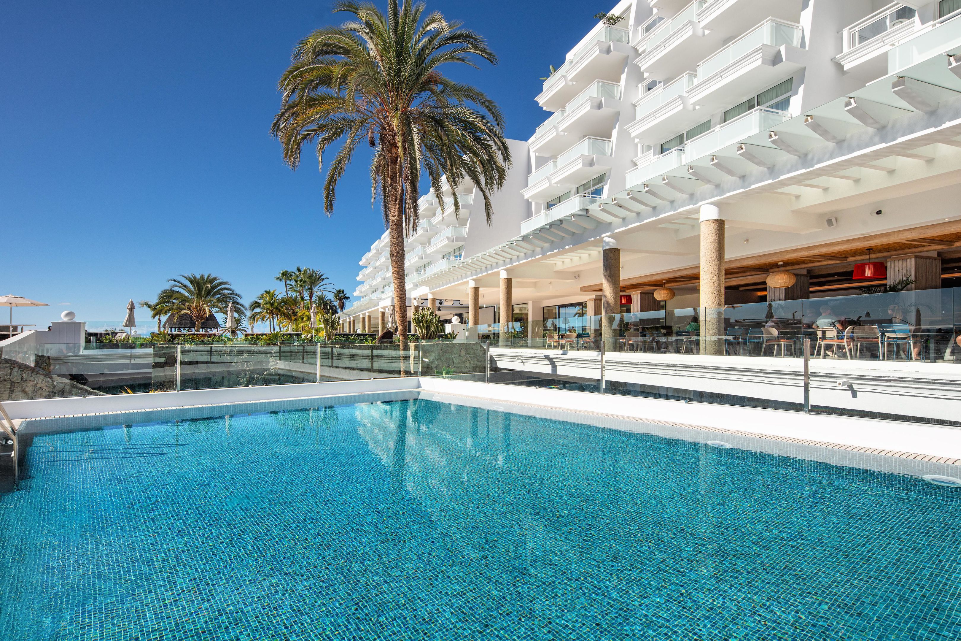 A swimming pool with clear blue water, surrounded by a white building with balconies and palm trees.