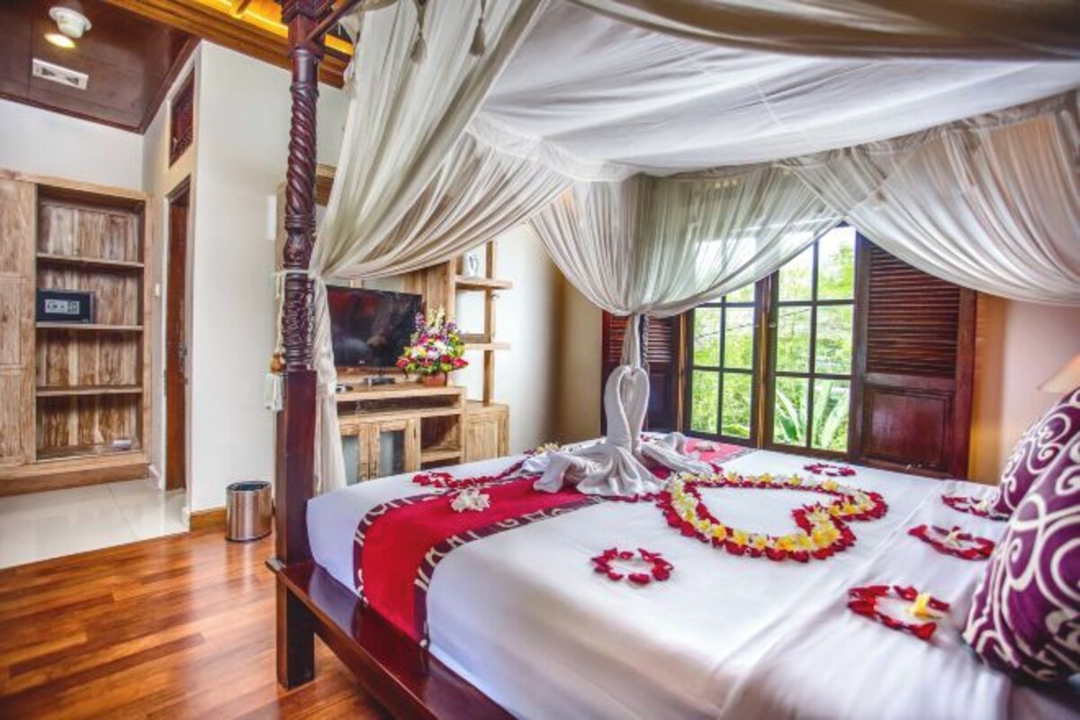 A four-poster bed with a canopy, a wooden cabinet, a television, and a window with a view of greenery.