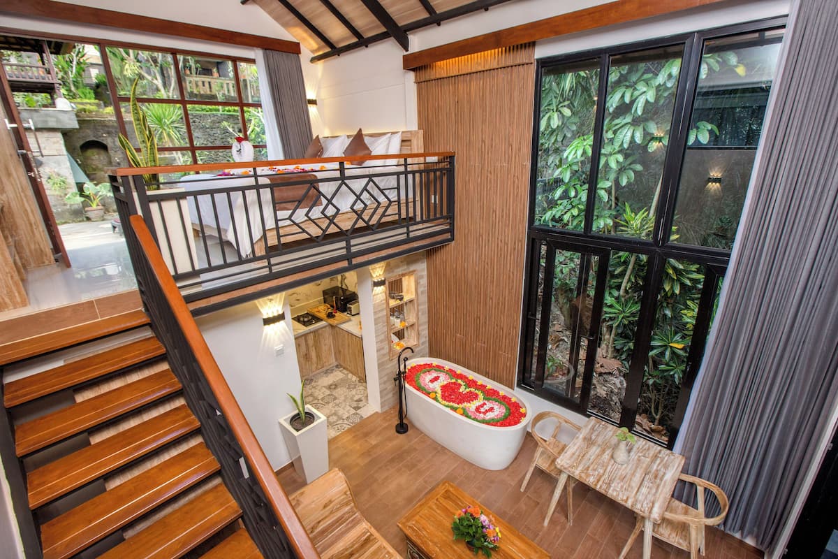 Un intérieur moderne avec un escalier en bois, un balcon doté d’une balustrade, une baignoire agrémentée d’un arrangement floral et de grandes fenêtres offrant une vue sur la verdure.