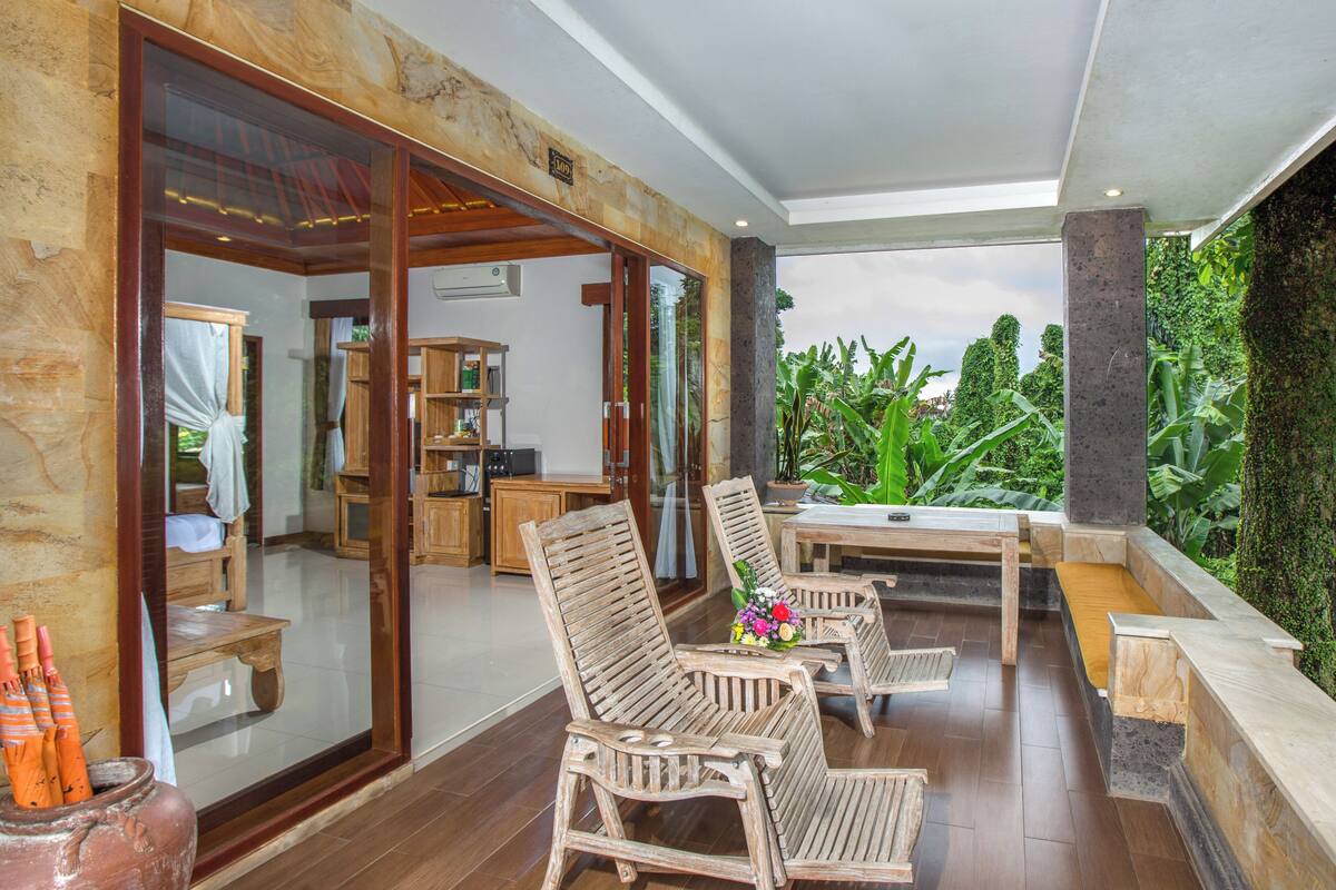 Um deck de madeira com mobiliário externo, vista para vegetação exuberante e uma porta de vidro que dá para o interior.