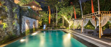 Una piscina con una pared de piedra y un pequeño puente, rodeada de sillones de descanso y telas colgantes.