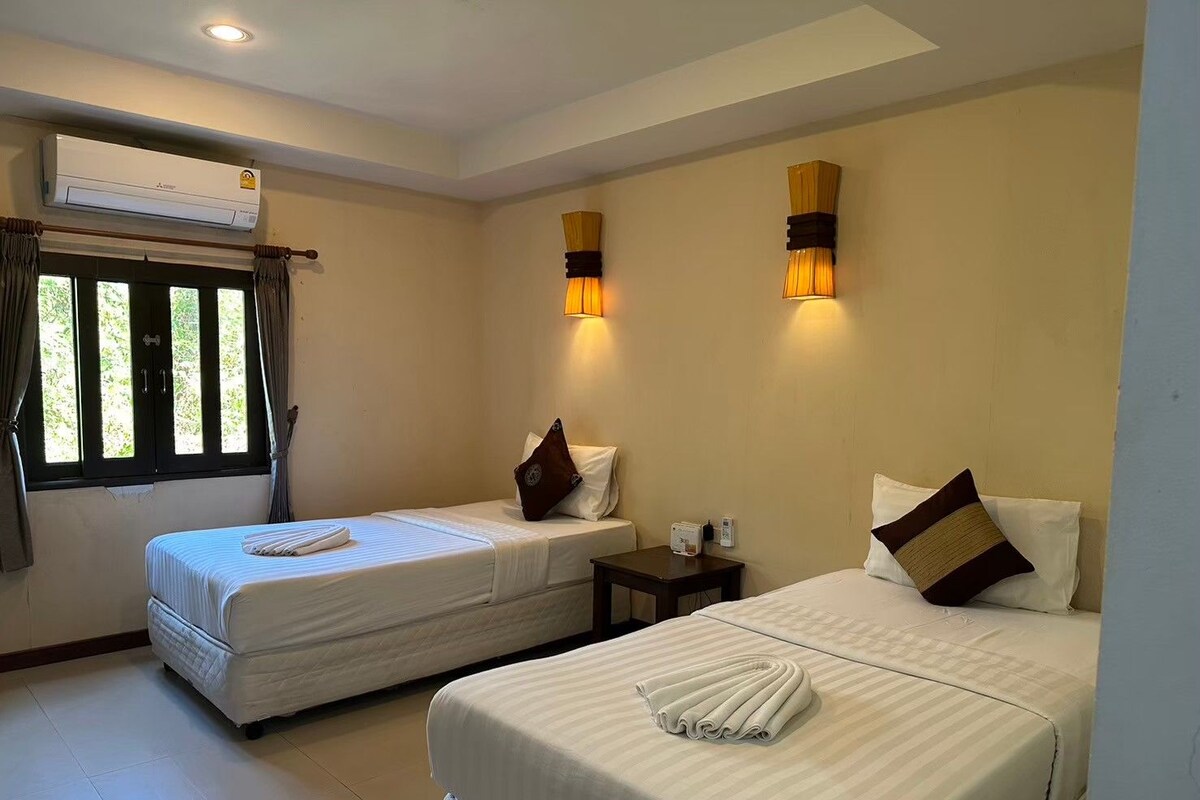 A hotel room with two beds, an air conditioning unit, a window with curtains, and wall-mounted lamps.