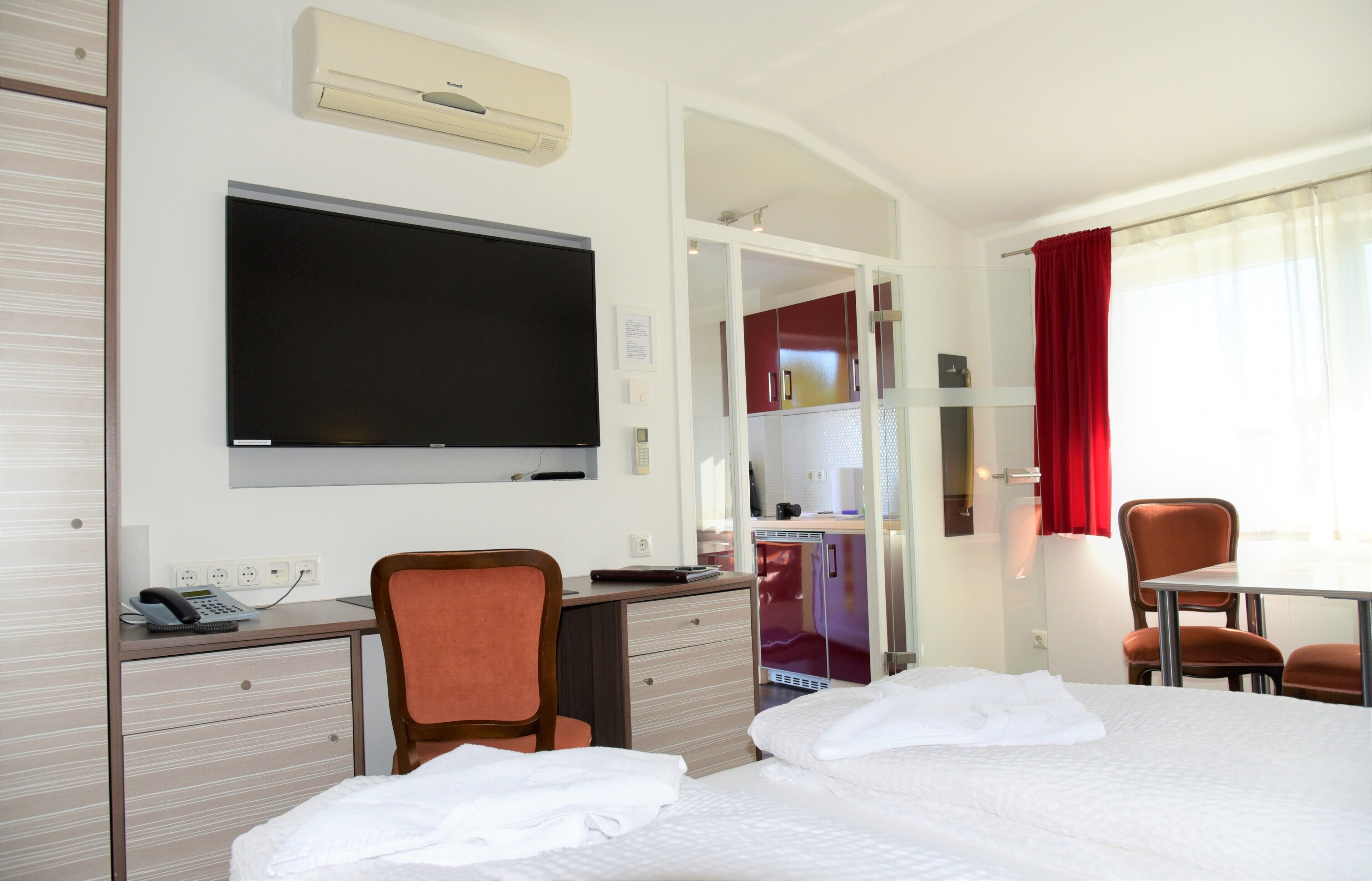 A hotel room with a bed, desk, chair, TV, and a small kitchenette.