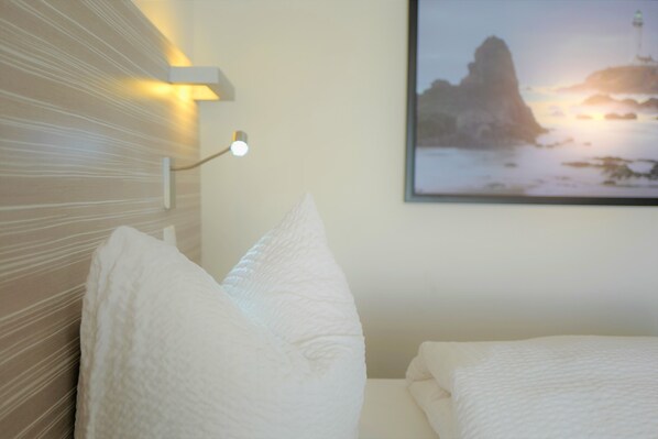 A bedroom with a bed, pillows, a wall-mounted light, and a framed picture of a coastal landscape.