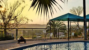 A pool area with chairs and a table under a gazebo, surrounded by a fence and palm trees.