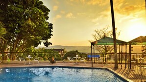 Una piscina con sillas de descanso y un pabellón, rodeada de árboles y vistas a montañas distantes.