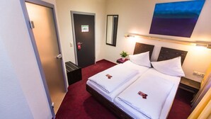 Habitación de hotel con dos camas, alfombra roja, mesita de noche con una planta, espejo y un cuadro en la pared.