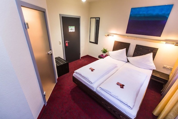 Une chambre d’hôtel avec deux lits, une moquette rouge, une table de chevet avec une plante, un miroir et un tableau au mur.