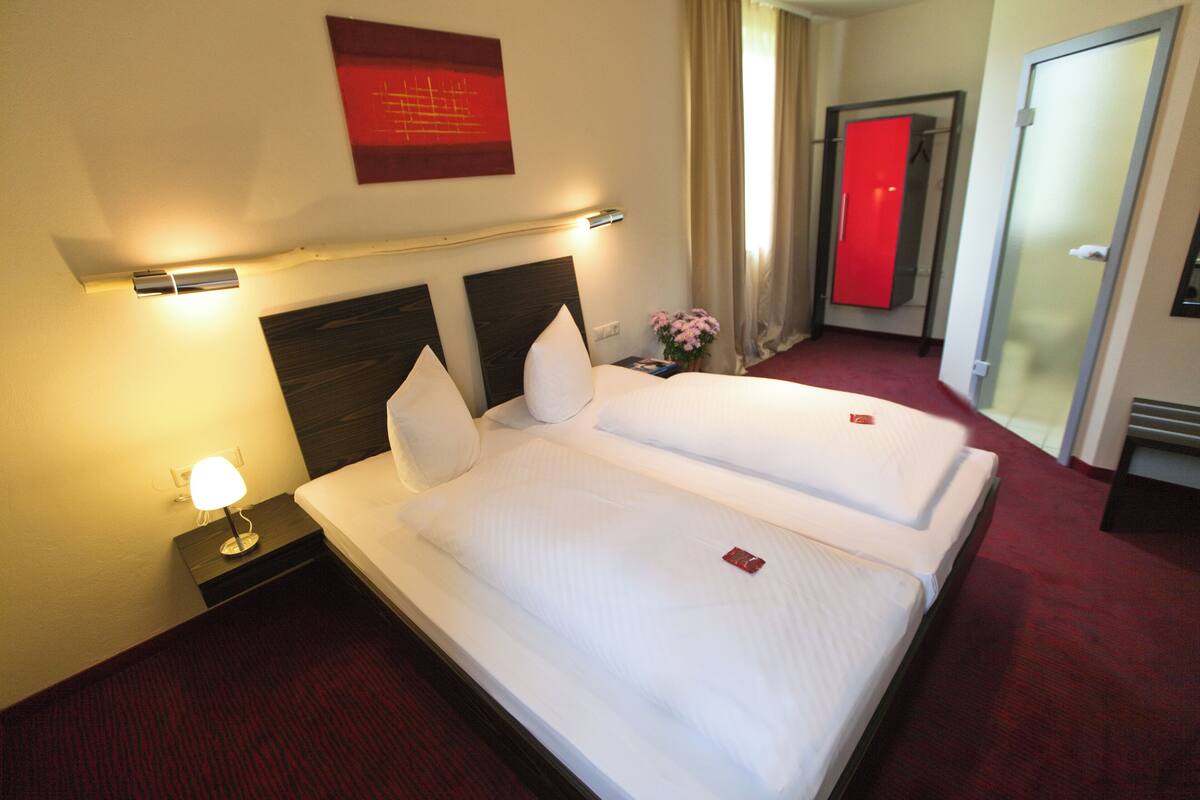 A hotel room with two beds, a nightstand with a lamp, a red door, and a red wall decoration.