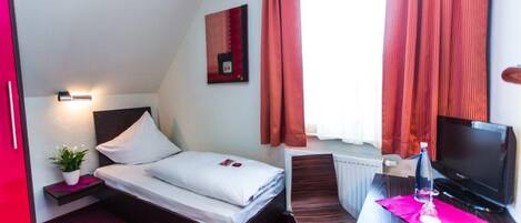 Habitación de hotel con cama, escritorio con televisor, silla, pequeña mesa con planta y ventana con cortinas.