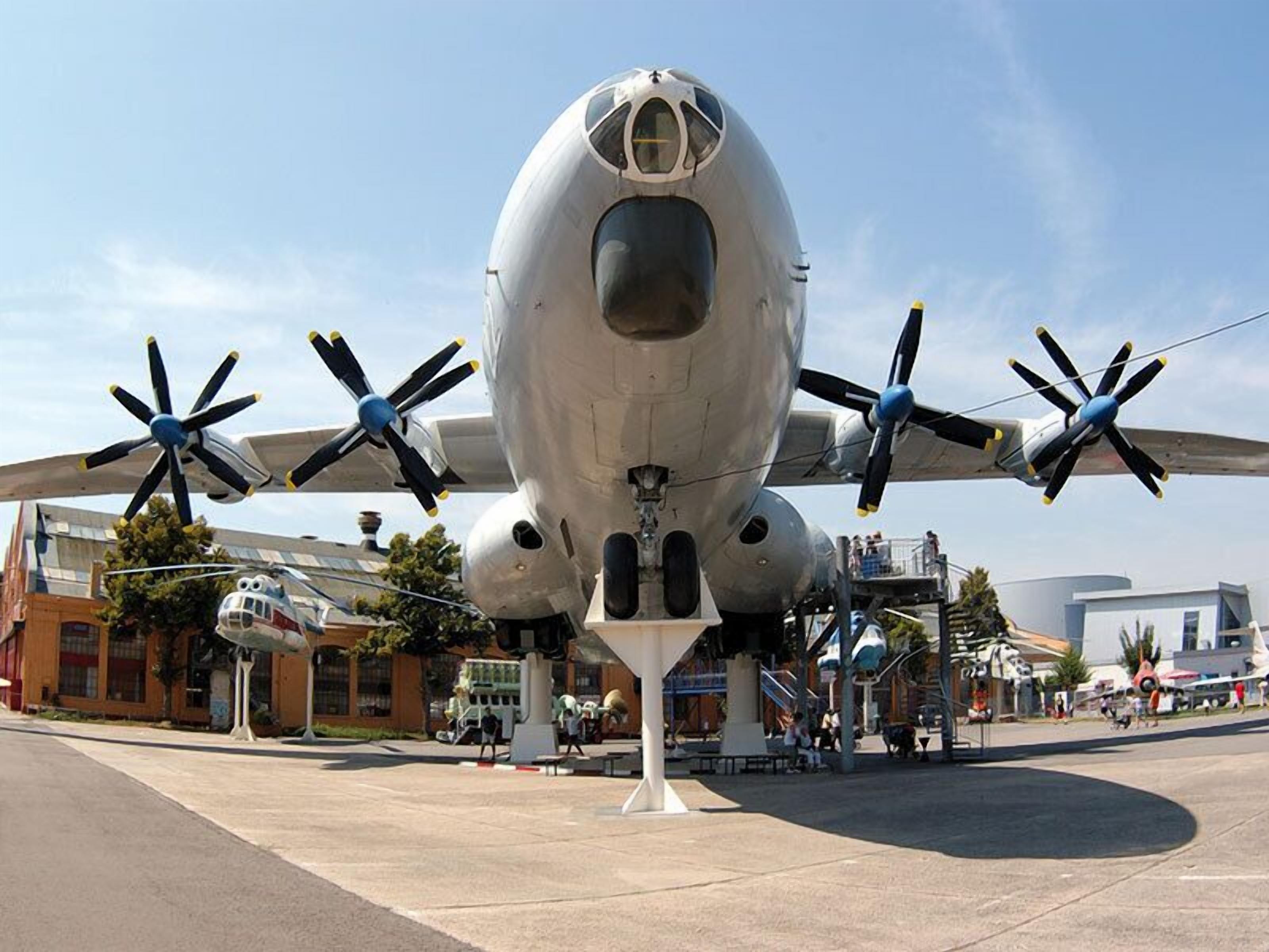 Una aeronave grande e inmóvil con cuatro hélices, probablemente una exhibición militar o histórica, rodeada de una zona pavimentada con edificios y gente al fondo.