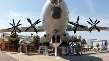 A large, stationary aircraft with four propellers, likely a military or historical exhibit, surrounded by a paved area with buildings and people in the background.