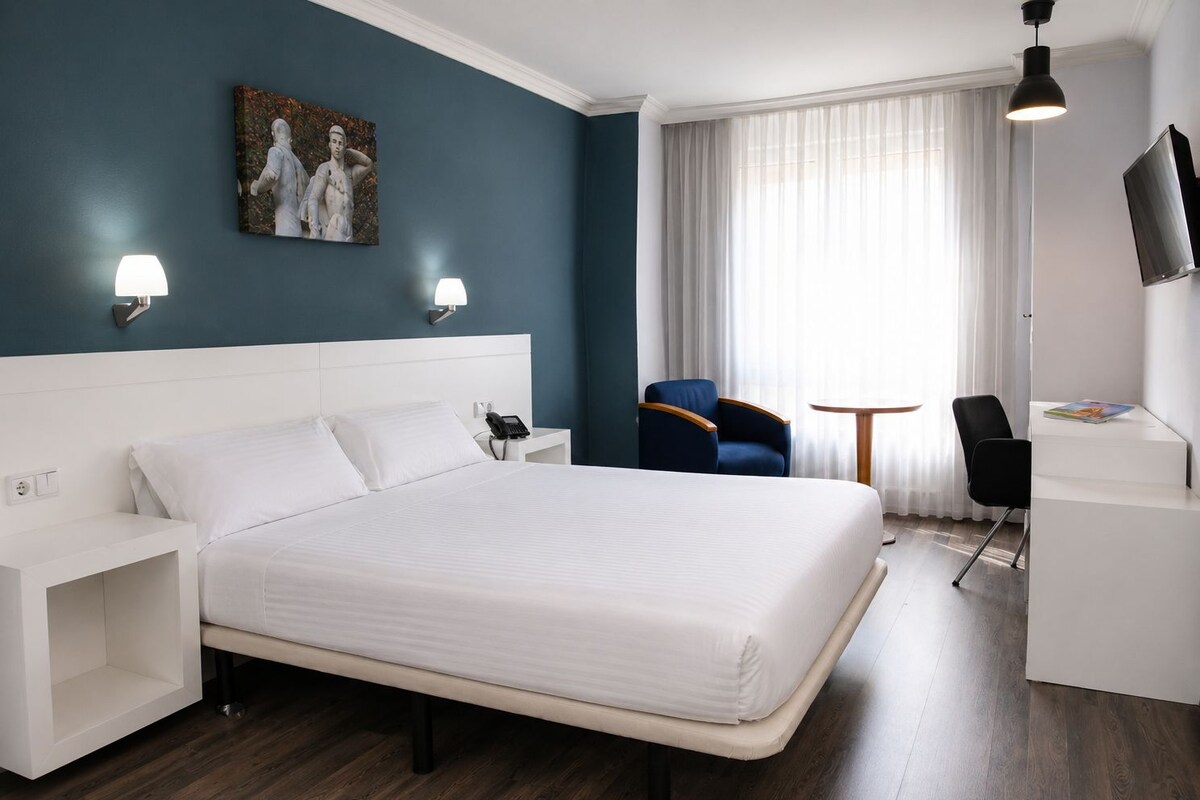 A hotel room with a large bed, a desk, a chair, a TV, and a painting on the wall.