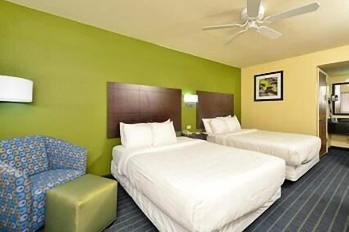 Quarto de hotel com duas camas, uma parede verde, um ventilador de teto e uma poltrona estampada.