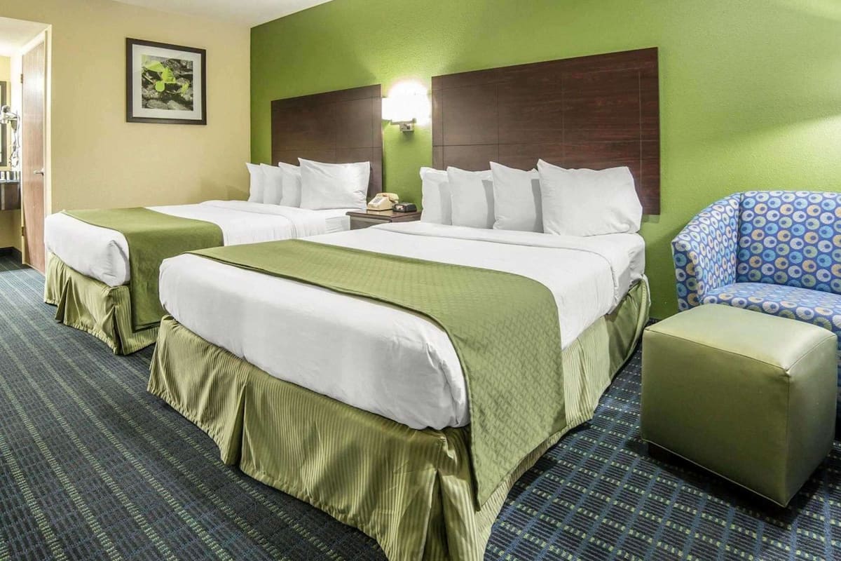 Quarto de hotel com duas camas, uma parede verde, ventoinha no teto, quadro emoldurado e poltrona estampada.