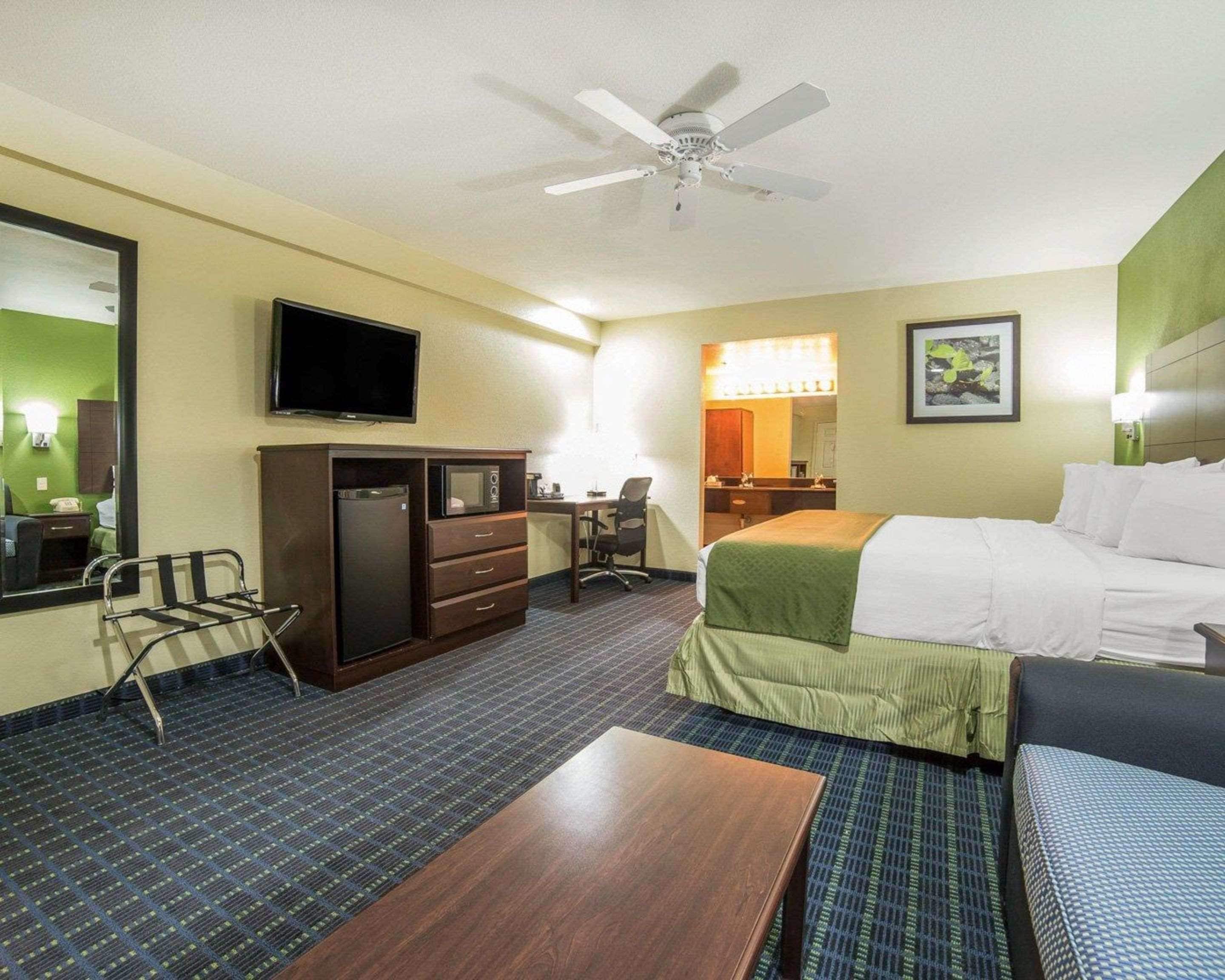 Hotelkamer met een bed, bureau, stoel, televisie, plafondventilator en een badkamer zichtbaar door een geopende deur.