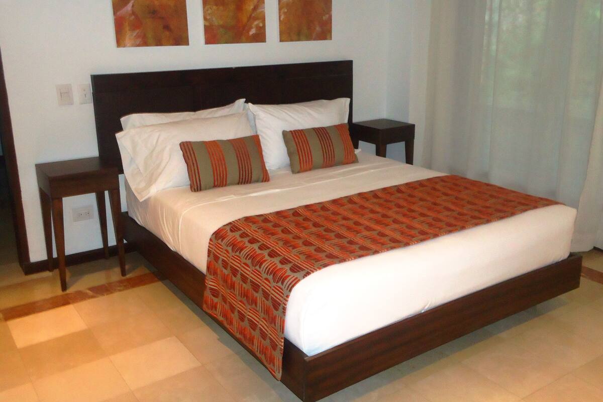 Una cama grande con estructura de madera, ropa de cama blanca y una manta decorativa estampada. Hay dos mesitas de noche, una a cada lado de la cama.