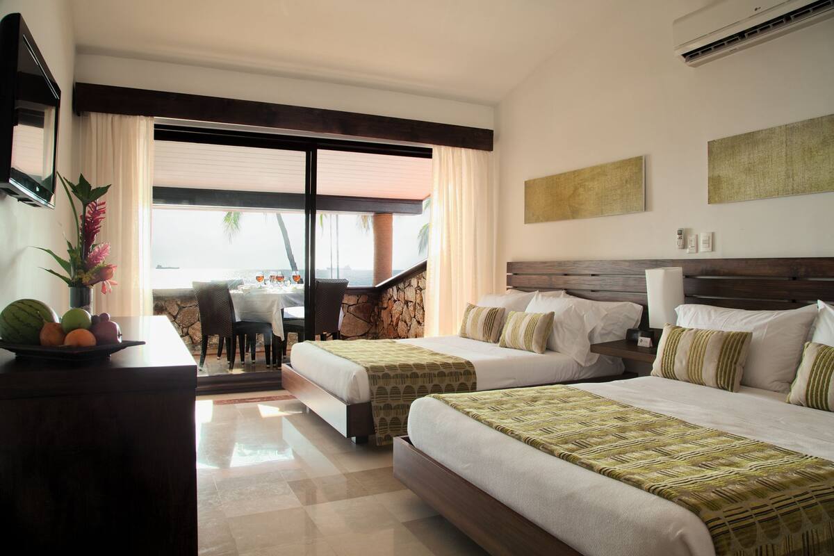 Habitación de hotel con dos camas, un comedor con mesa y sillas, y vista al mar.
