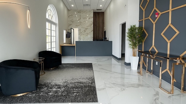 A modern lobby with a reception desk, a black sofa, a potted plant, and a geometric wall design.