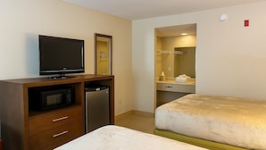 A hotel room with a bed, a TV, a mini-fridge, a wooden dresser, a bathroom with a sink and mirror, and a red emergency light.
