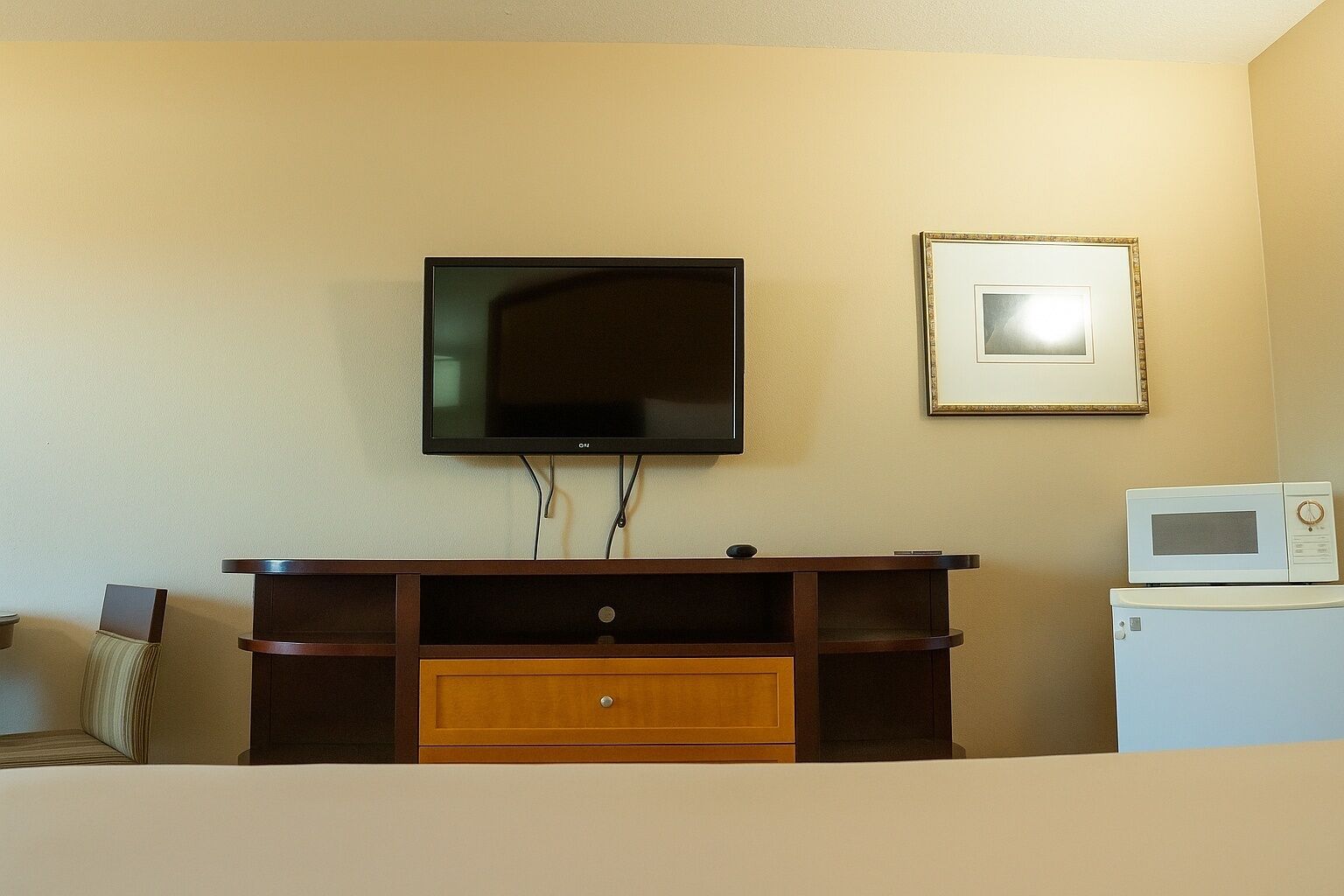 Une pièce avec un téléviseur fixé au mur, un tableau encadré, un meuble en bois avec des tiroirs et un micro-ondes posé sur une petite table.