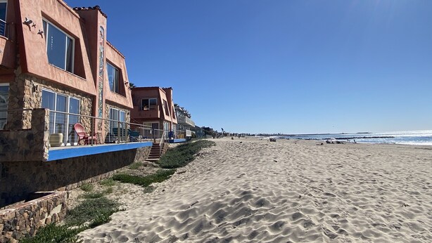 A beachfront hotel with stone and stucco exteriors, large windows, and a blue railing along the beach. - The Shores Inn and Beach Houses (Ventura)