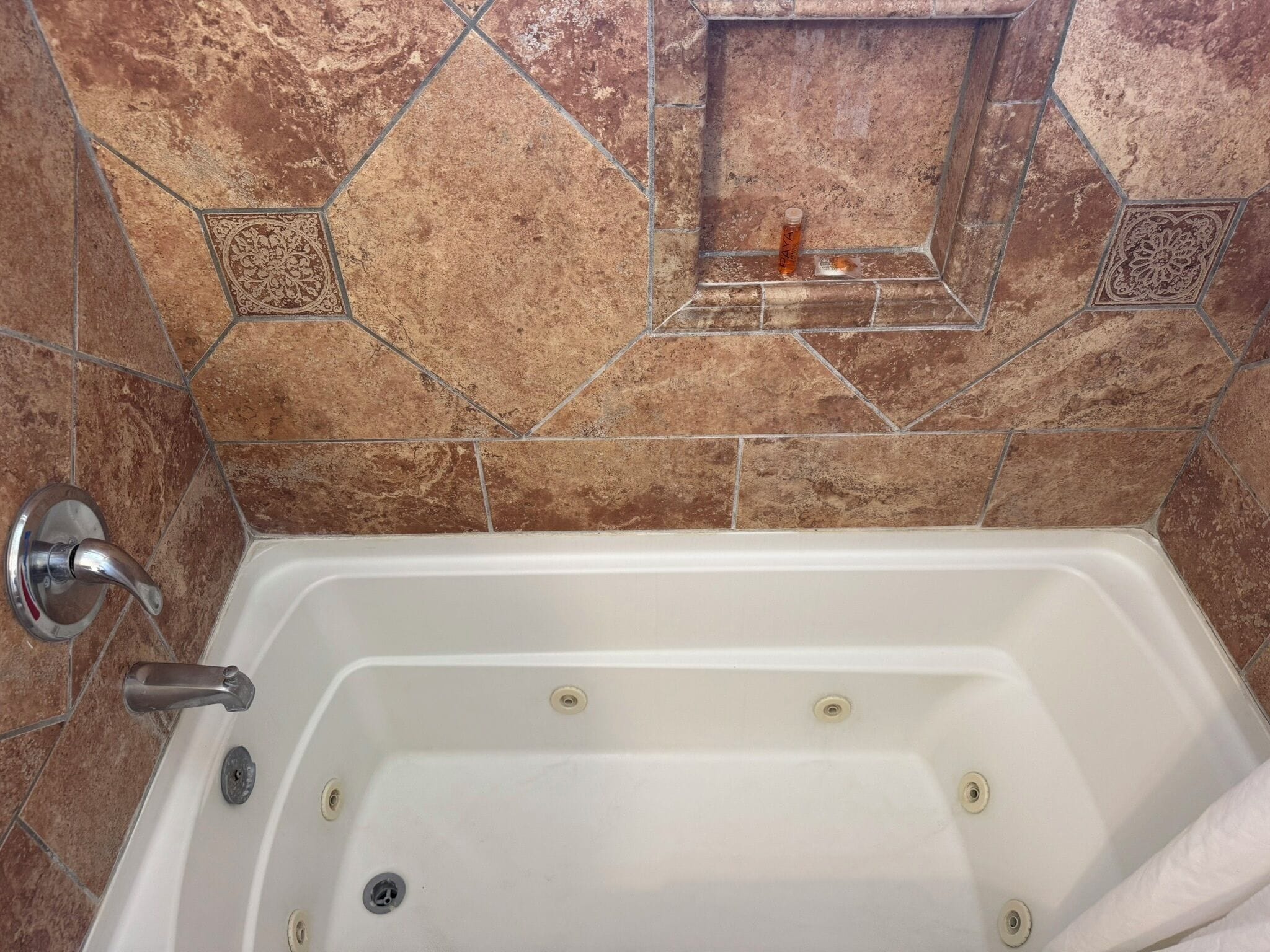 Eine Badewanne mit Duschkopf, eine kleine Ablagefläche mit einer Flasche und eine Wand mit dekorativen Fliesen.