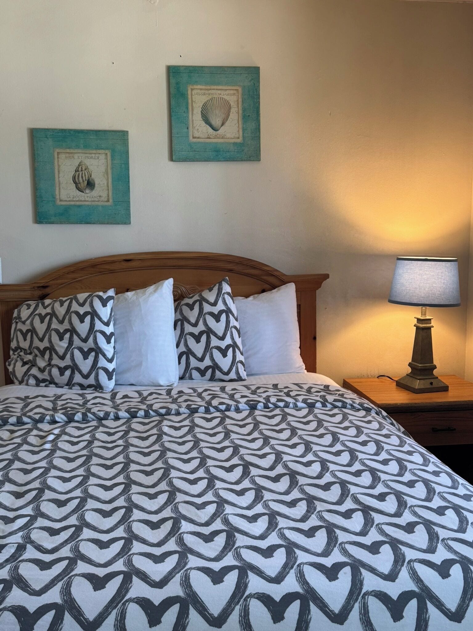 A bed with a patterned bedspread, two heart-shaped pillows, a wooden headboard, a nightstand with a lamp, and two framed pictures on the wall.