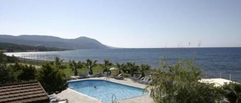 A pool area with lounge chairs and a small building, overlooking a body of water and hills.