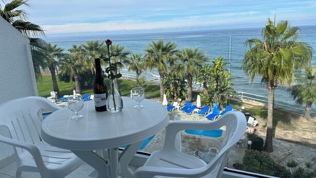 Un balcon avec une table dressée pour deux personnes, donnant sur une plage bordée de palmiers et offrant une vue sur l’océan.