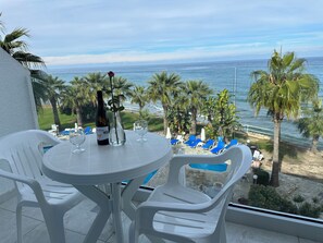 A balcony with a table set for two, overlooking a beach with palm trees and a view of the ocean.